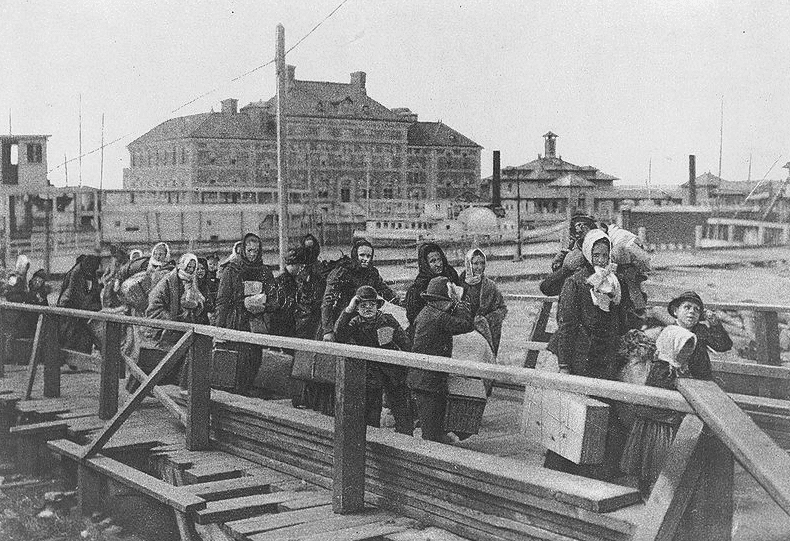 Photo: “Emigrants coming up the board-walk from the barge, which has taken them off the steamship company’s docks, and transported them to Ellis Island. The big building in the background is the new hospital just opened,” 1902
